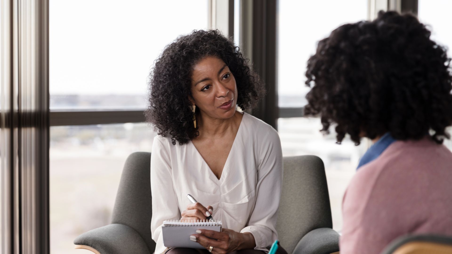 Two women seated in armchairs engage in a serious conversation, with one woman attentively taking notes while the other is speaking.
